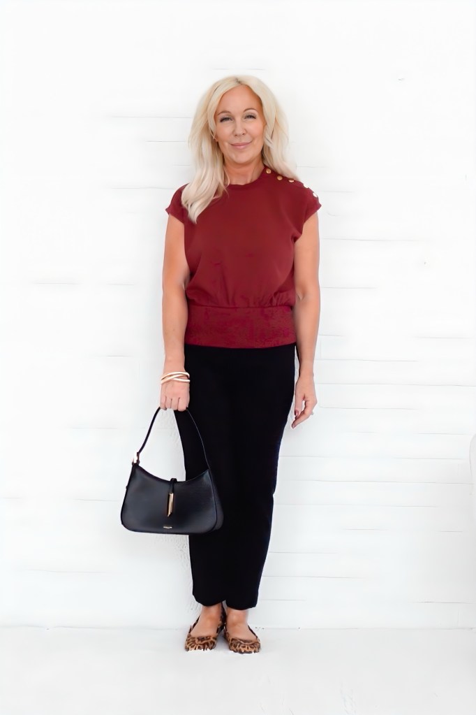 A woman with long, blonde hair wearing a burgundy top and black skirt holds a black handbag while standing against a white background.