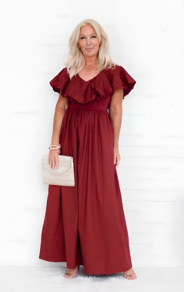 A woman wearing a burgundy maxi dress with ruffled sleeves, holding a light-colored clutch, standing against a white background.