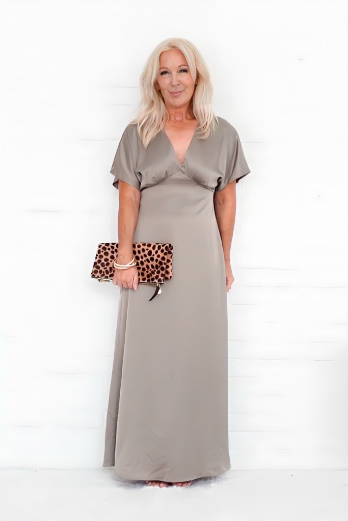 A woman in a flowing beige dress holds a leopard print clutch, standing against a white background.