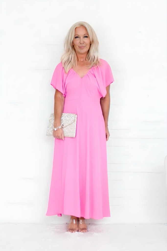 A woman wearing a flowing pink dress with short sleeves, holding a silver clutch, standing in front of a white background.