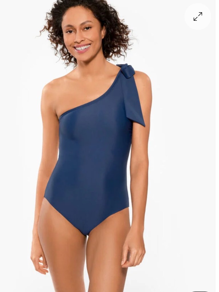 A woman with curly hair smiles while wearing a one-shoulder navy blue swimsuit with a bow detail on the shoulder.