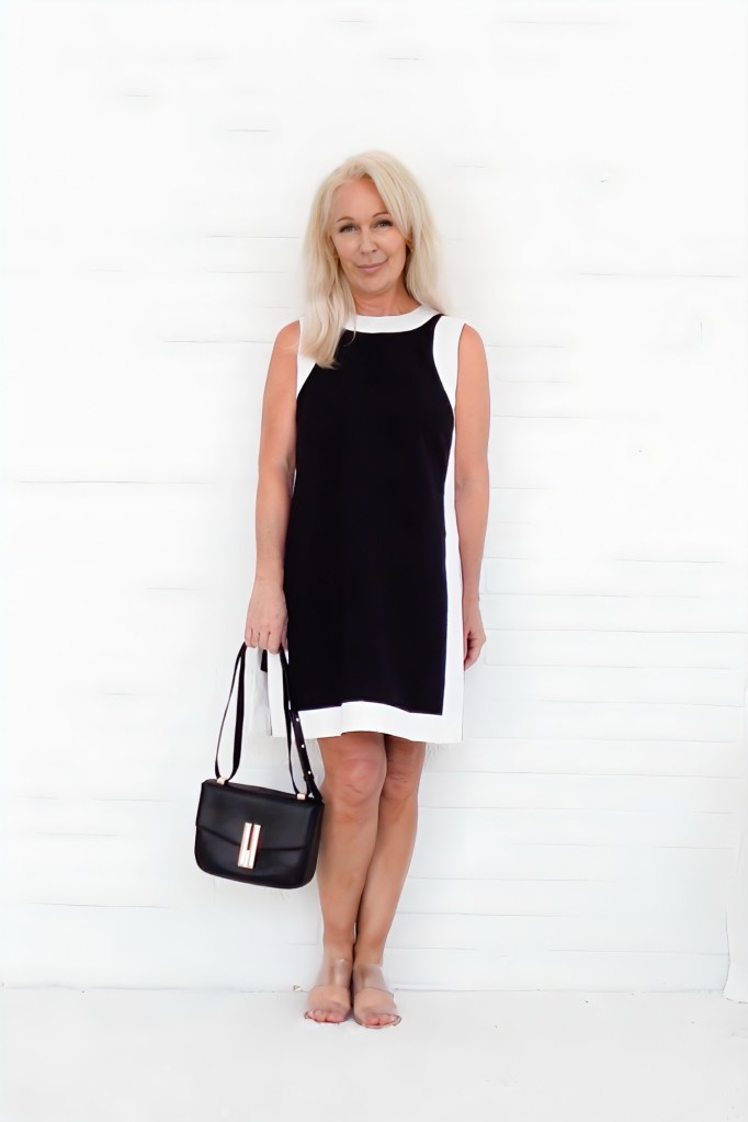 A woman wearing a black and white dress, holding a black handbag, standing against a white backdrop.