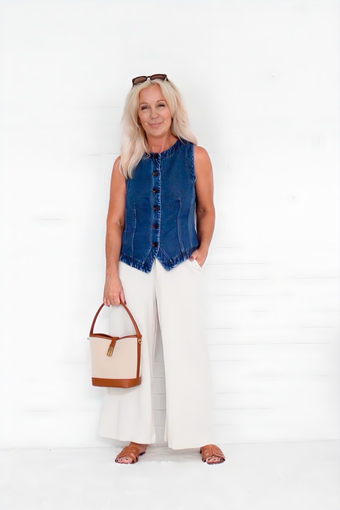 A woman standing against a white background, wearing a denim vest, wide-leg cream pants, and brown sandals, holding a small handbag.