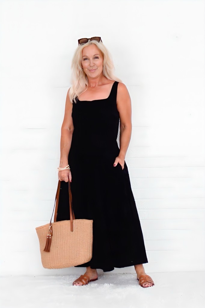 A woman with long blonde hair wearing a black summer dress stands against a white background, holding a woven tote bag with brown straps.