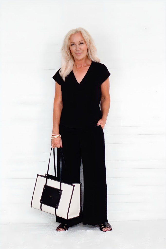 A woman with long light hair wearing a black outfit and holding a large tote bag, standing against a white background.
