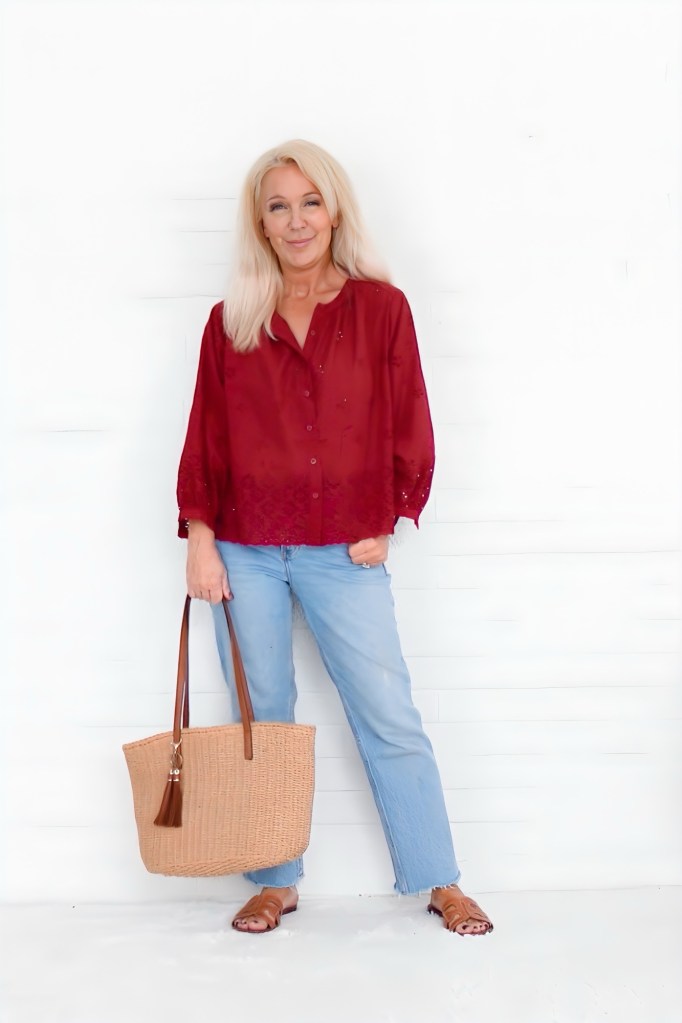 A woman with long blonde hair wearing a burgundy button-up blouse, relaxed light-wash jeans, and brown slip-on sandals, holding a woven tote bag, standing against a white backdrop.