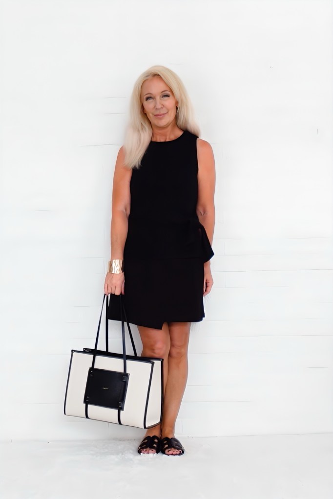 A woman wearing a black sleeveless dress and holding a large black and white tote bag, standing against a white background.