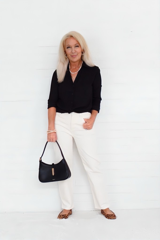 A woman with long blonde hair wearing a black button-up shirt and ivory pants, posing with a black handbag and leopard-print flats against a white background, exuding a stylish and polished look.