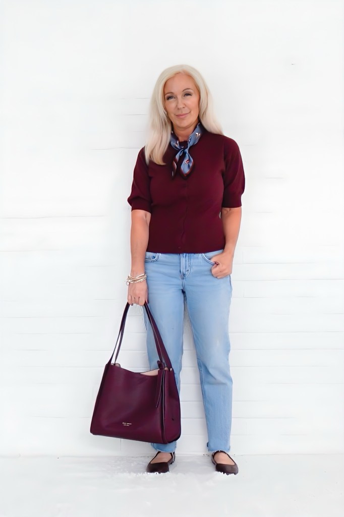 A woman stands in front of a white background, wearing a maroon top, light blue jeans, and black flats. She holds a burgundy handbag and has a blue scarf around her neck, sporting a friendly smile.