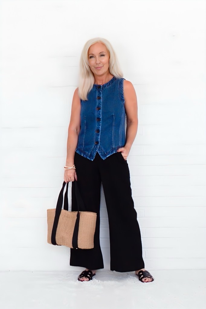 A woman with long blonde hair wearing a sleeveless denim vest and black wide-leg pants, standing against a white background, holding a woven straw bag.