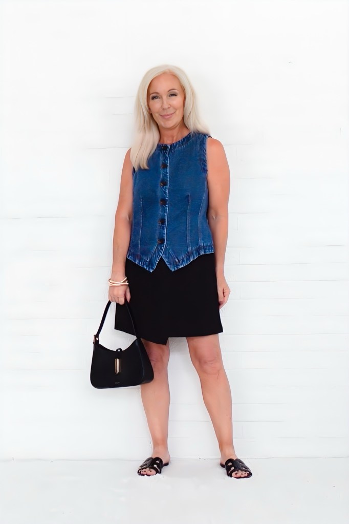 A woman with long blonde hair is wearing a sleeveless blue denim vest paired with a black skirt, holding a black handbag in front of a white background.