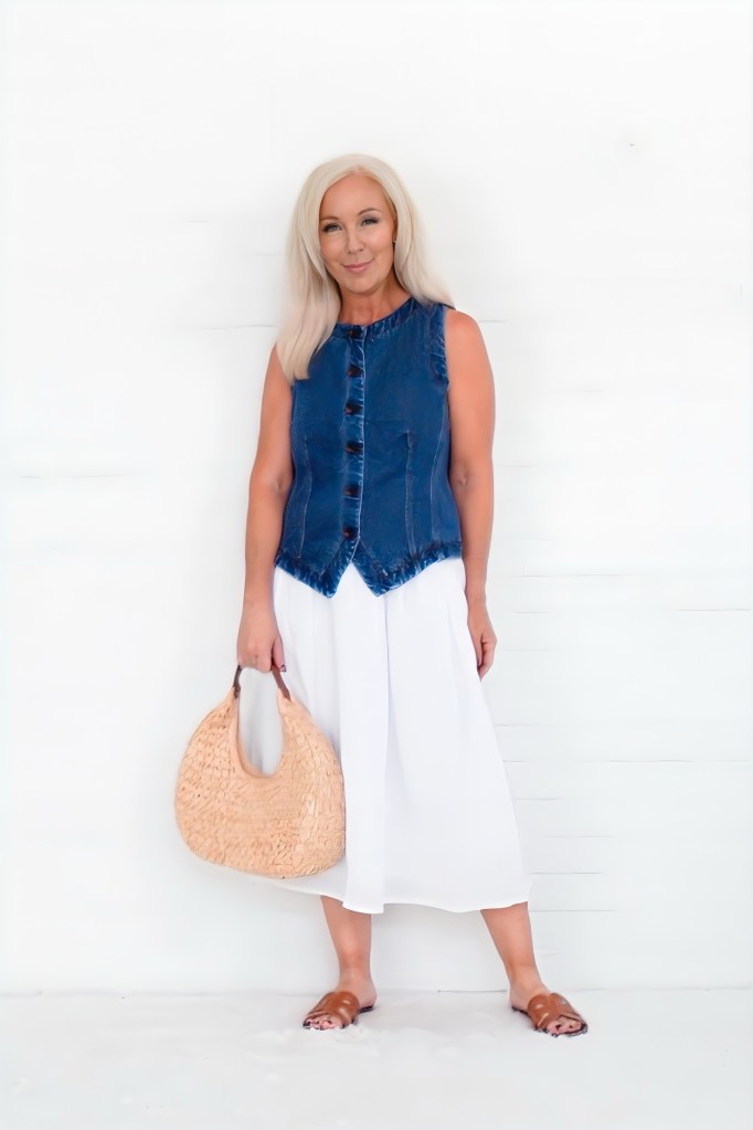 A woman with long blonde hair wearing a sleeveless denim vest and a white flowy midi skirt, holding a woven straw bag.