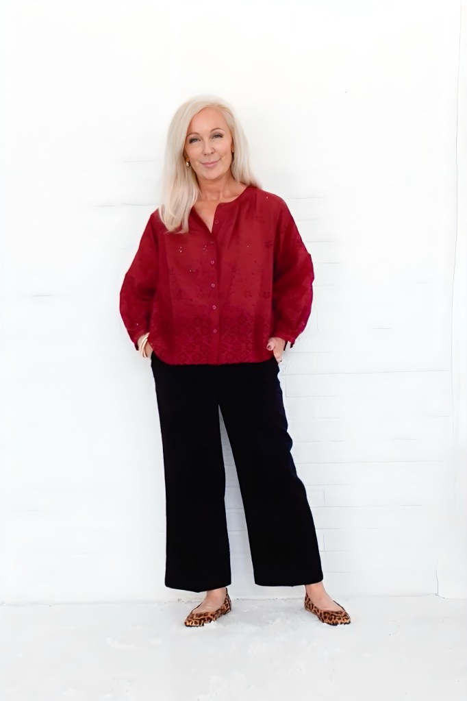 A woman with long, straight white hair smiles confidently while wearing a red button-up top, black trousers, and leopard print flats, standing against a white background.