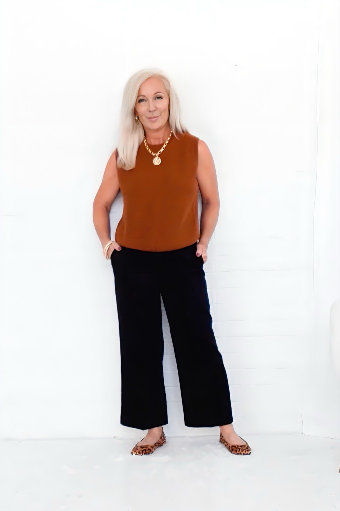 A mature woman with blonde hair stands confidently against a white wall, wearing a brown sleeveless top, black wide-leg pants, and leopard print flats, accessorized with a gold necklace.