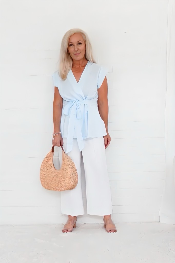 A woman stands confidently against a white background, wearing a light blue wrap top and white wide-leg trousers, holding a woven straw bag.