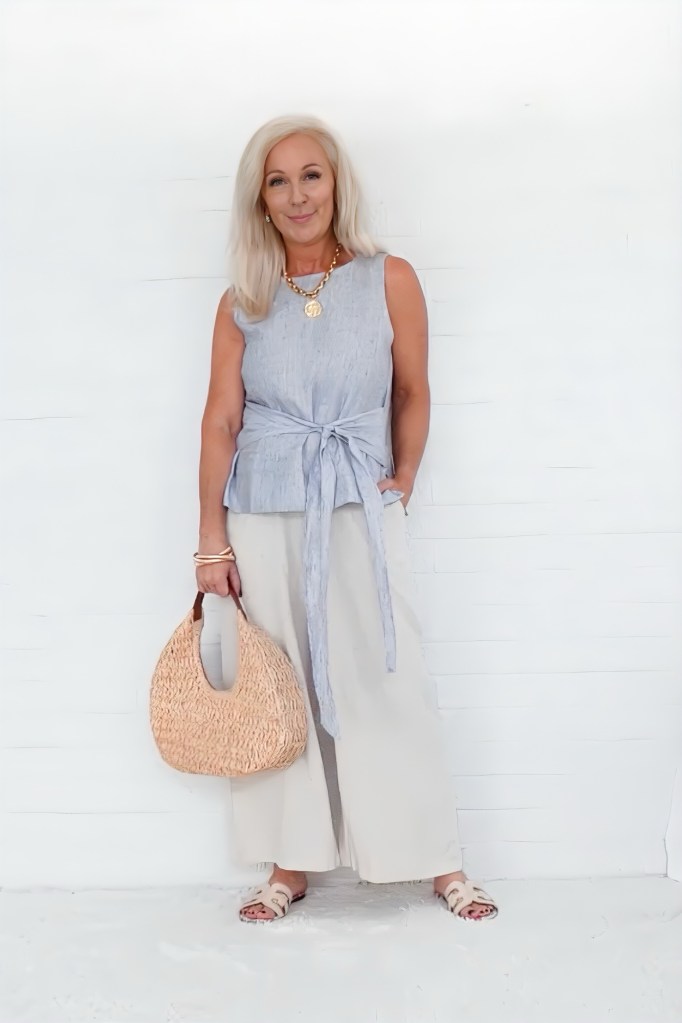 A woman stands confidently in a stylish outfit, featuring a sleeveless top with a tied detail and wide-leg pants, holding a woven bag and smiling against a plain white background.
