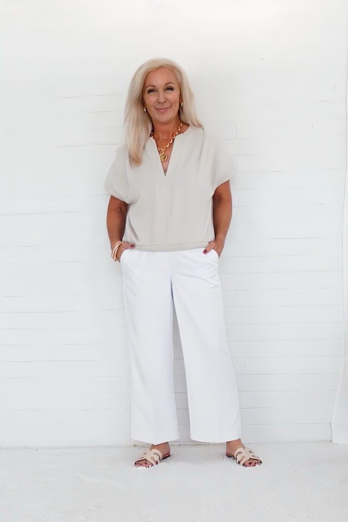 A smiling woman with blonde hair stands against a white backdrop, wearing a light gray blouse and white wide-leg pants, with her hands in the pockets and gold jewelry accessorizing her outfit.