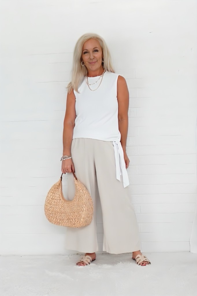 A woman with long, light hair wearing a white sleeveless top and beige wide-leg pants, holding a straw bag, standing against a white wall.