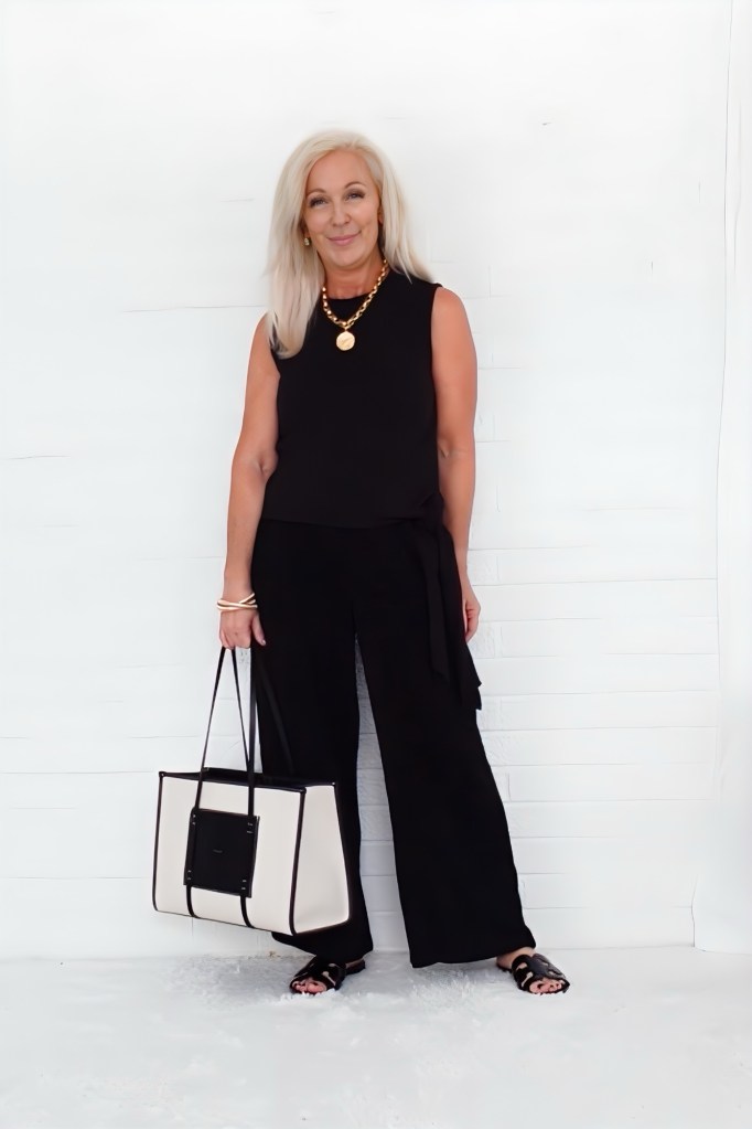 A woman with long light hair smiles while wearing a sleeveless black top and wide-leg black pants, holding a stylish black-and-white handbag against a plain white background.