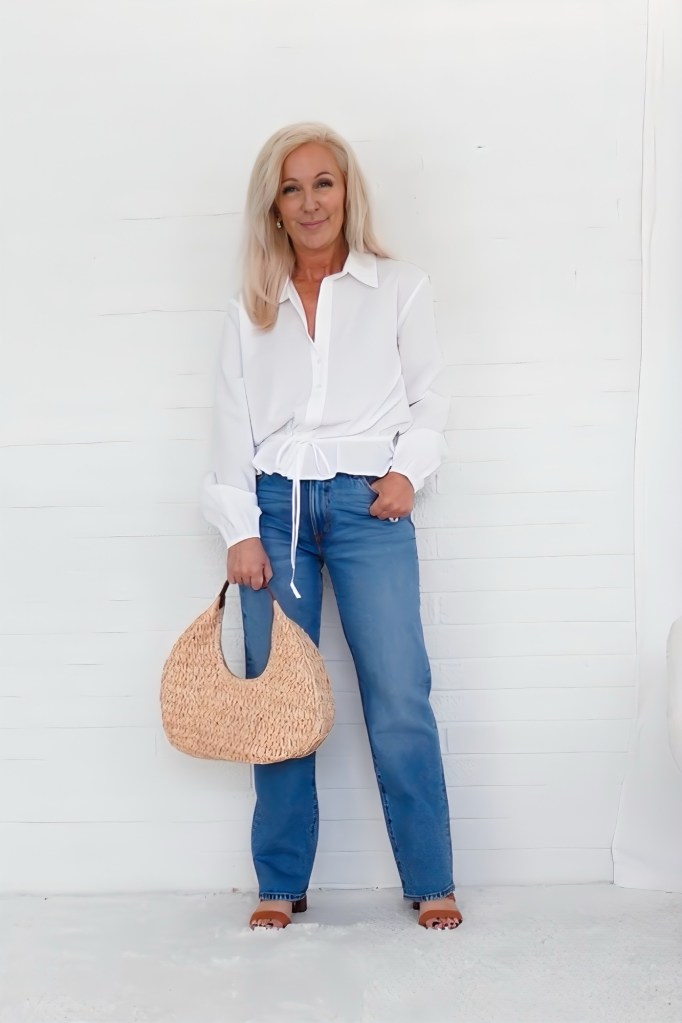 A woman with long blonde hair wearing a white button-up shirt and blue jeans, holding a woven bag, standing against a white wall.