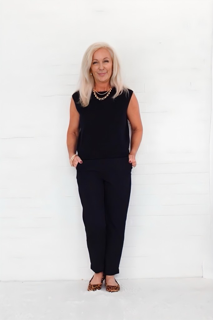 A confident woman standing in a stylish black outfit with a relaxed fit, featuring a sleeveless top and pants, accessorized with a necklace, against a plain white background.