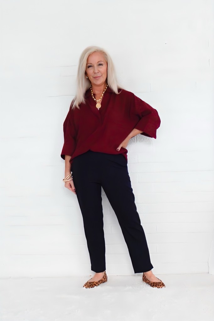 A stylish older woman stands confidently against a white background, wearing a burgundy blouse and black pants, paired with leopard print flats and accessorized with jewelry.