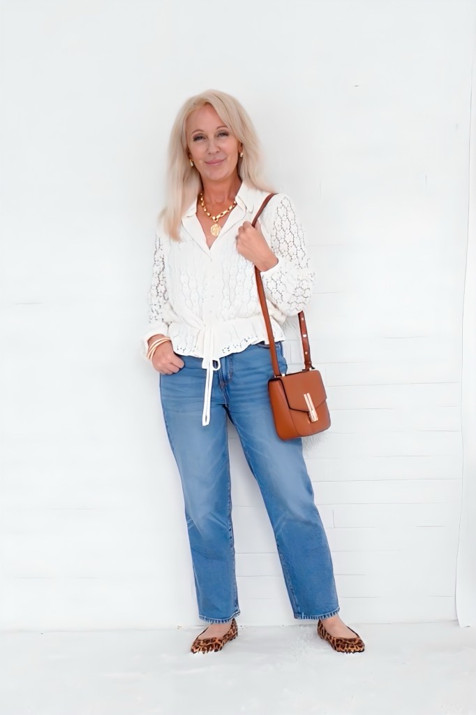 A stylish woman stands against a white background, wearing a white lace top, light blue jeans, and leopard print shoes. She holds a brown crossbody bag and smiles confidently, accessorized with layered necklaces.
