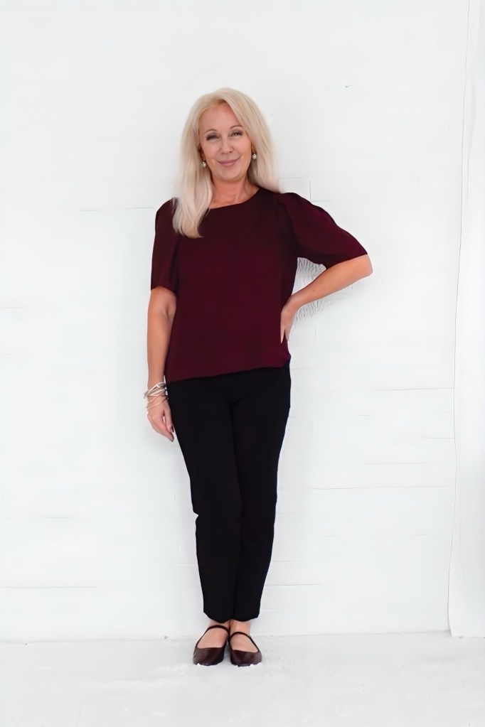 A woman smiling while standing against a white wall, wearing a burgundy top and black pants, with her hands on her hips and bracelets on her wrist.