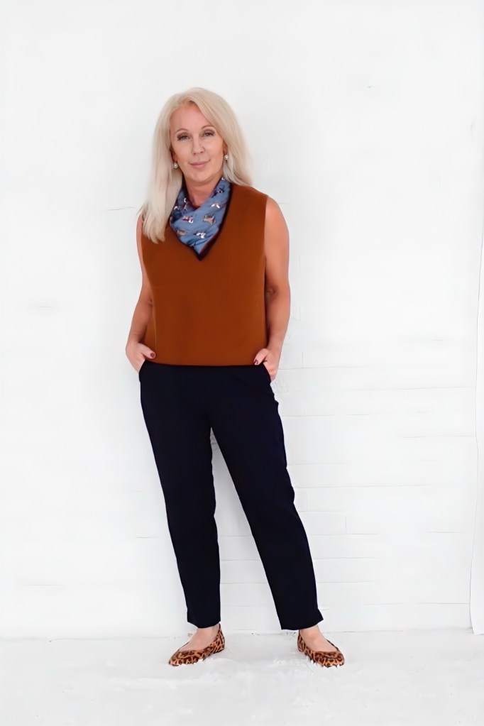 A woman with long blonde hair stands confidently against a white background, wearing a brown sleeveless top, navy pants, and leopard print flats, accessorized with a blue patterned scarf.
