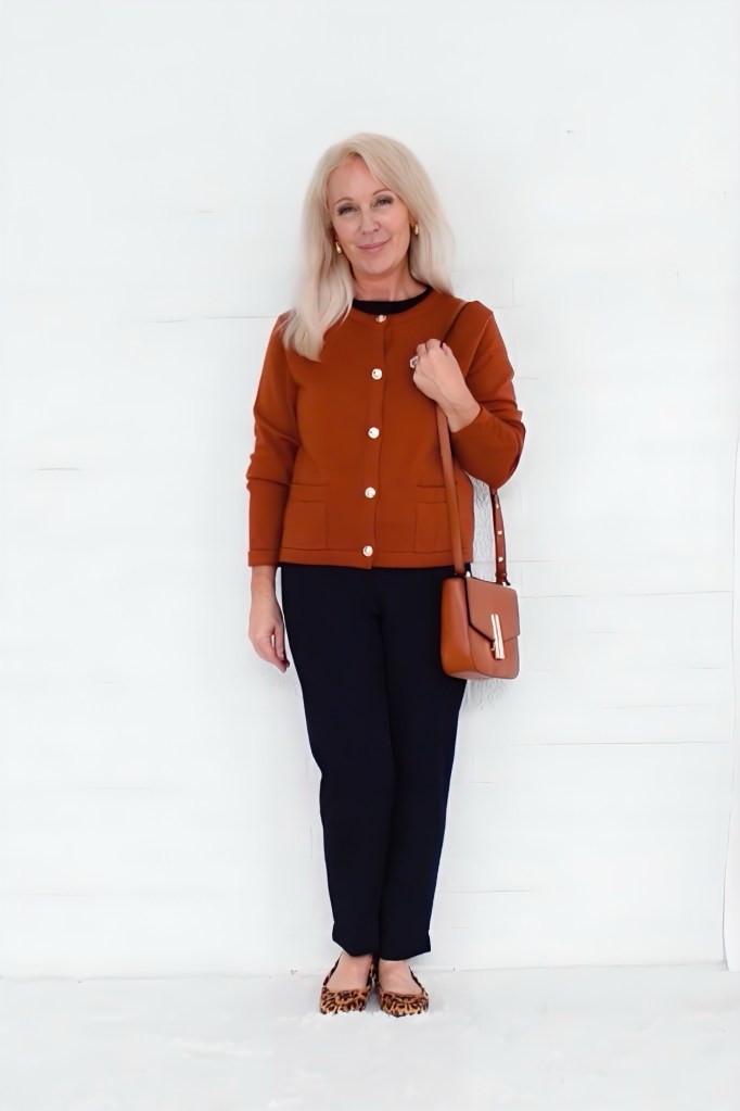 A woman with long blonde hair wearing a rust-colored cardigan, navy pants, and leopard print flats stands against a white background, holding a brown crossbody bag.