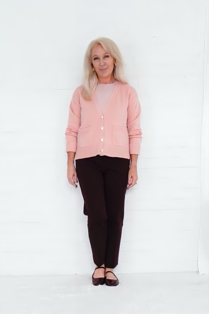 A woman standing against a white background, wearing a light pink cardigan over a matching top and dark brown pants, and smiling at the camera.
