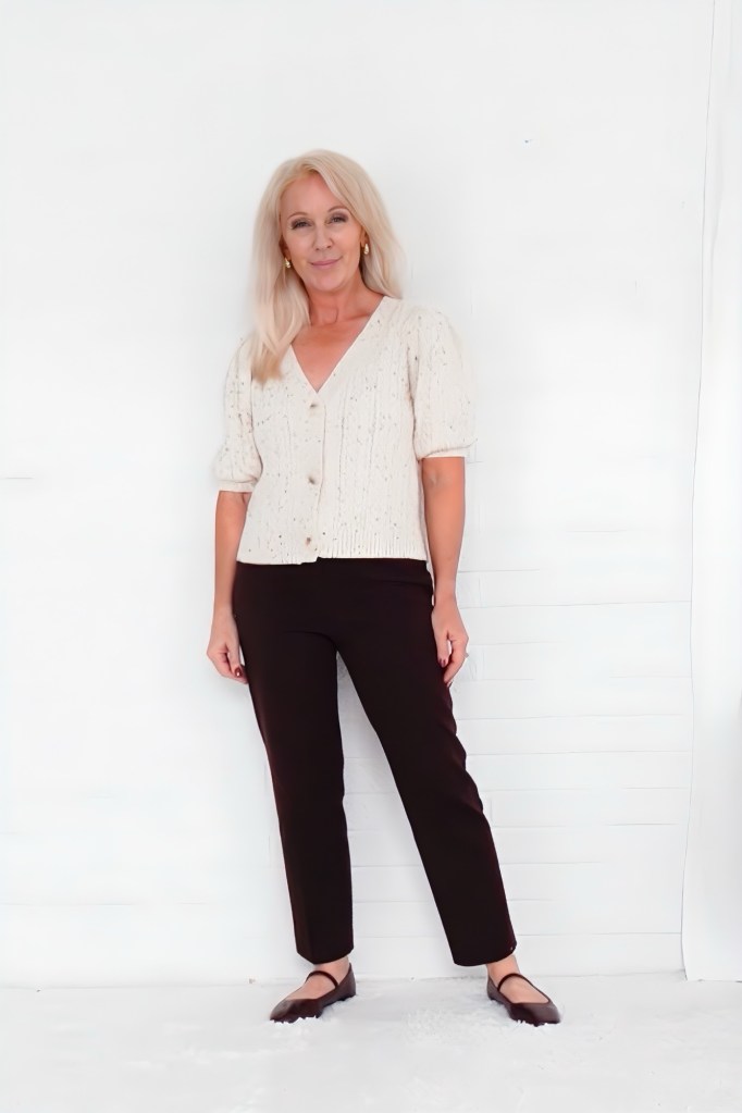 A woman with long blonde hair stands confidently against a white backdrop, wearing a light-colored, short-sleeved knit top and dark pants, exuding a stylish and approachable vibe.