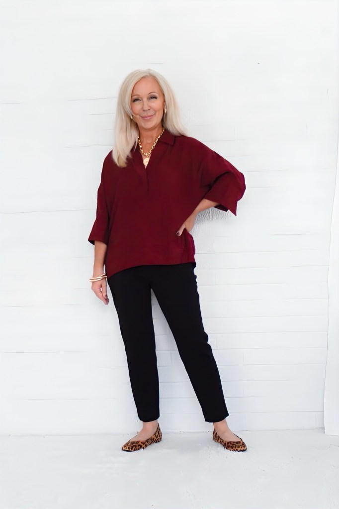 A woman with long blonde hair stands against a white background, wearing a burgundy top, black pants, and leopard print flats, smiling confidently.