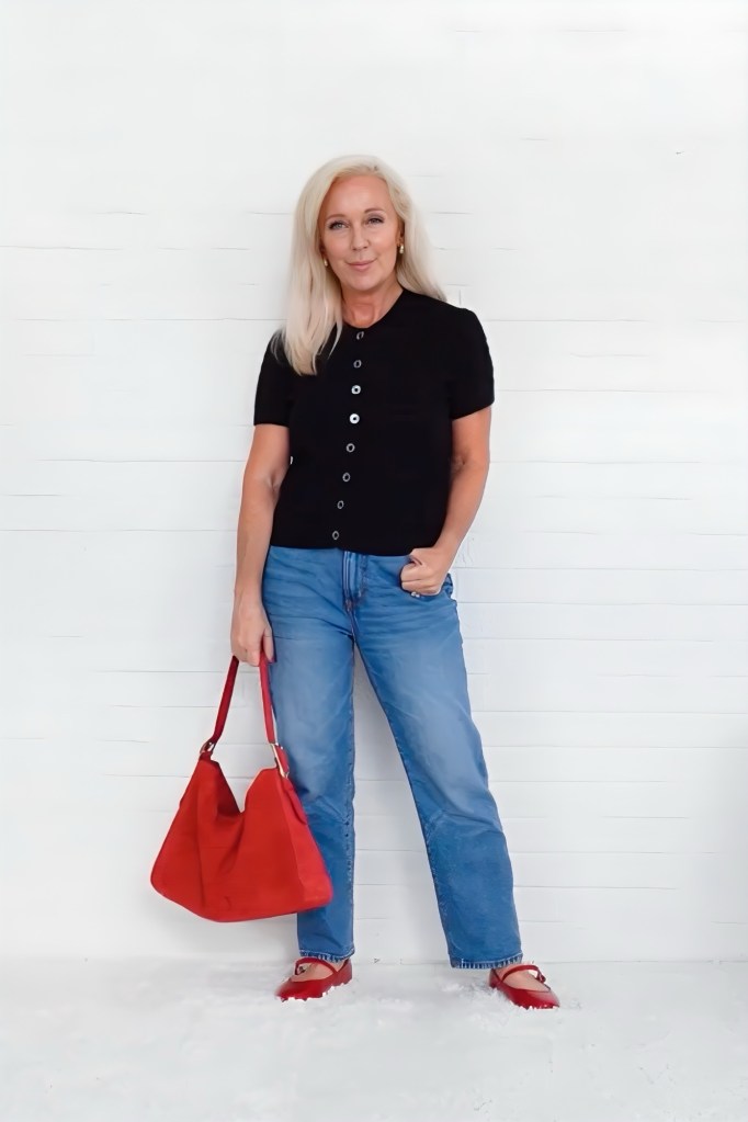A middle-aged woman stands against a white background, wearing a black short-sleeve top and blue straight leg jeans, holding a red handbag.