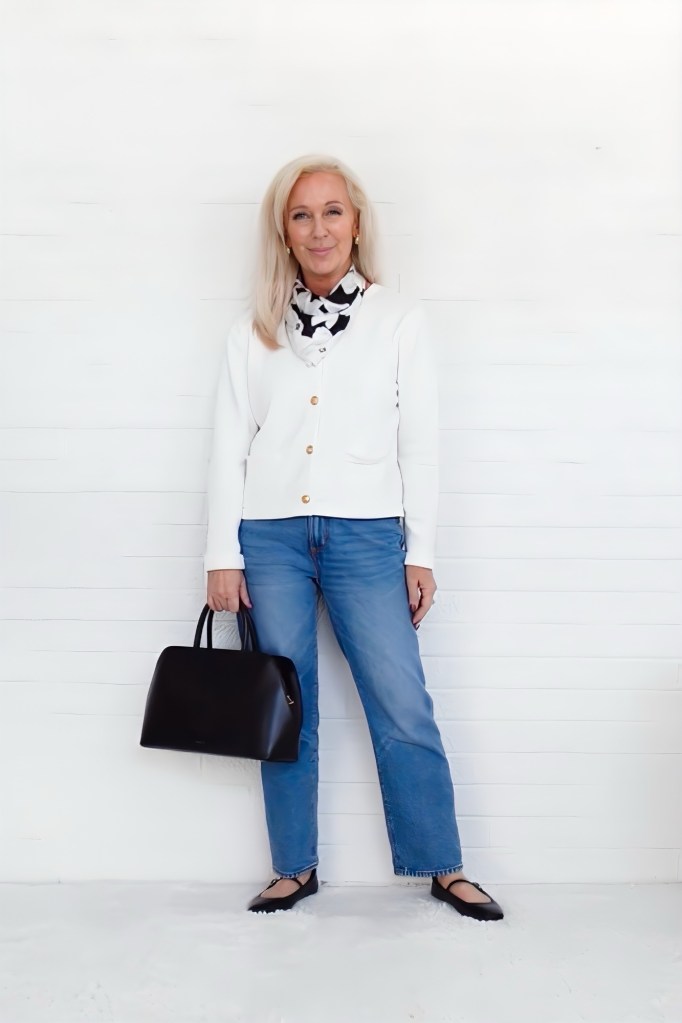A woman standing in front of a white wall, wearing a white cardigan over a patterned scarf, with blue jeans and black flats, holding a black handbag.