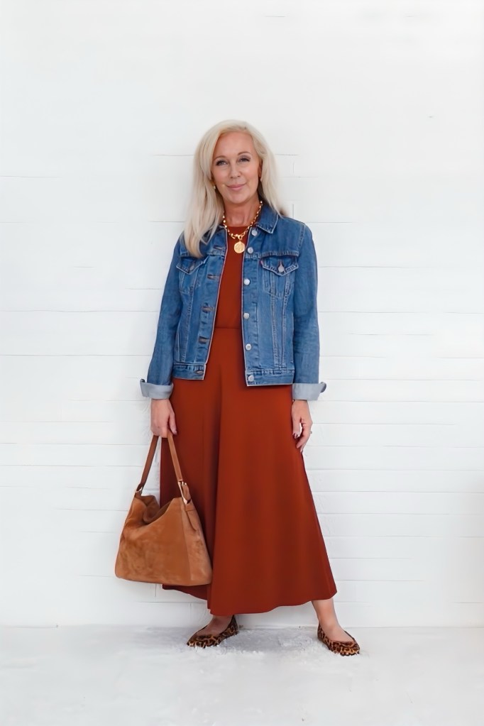 A woman with long, silver hair stands in front of a white wall, wearing a rust-colored midi dress paired with a denim jacket. She holds a tan handbag and wears leopard print flats, accessorized with layered gold necklaces.