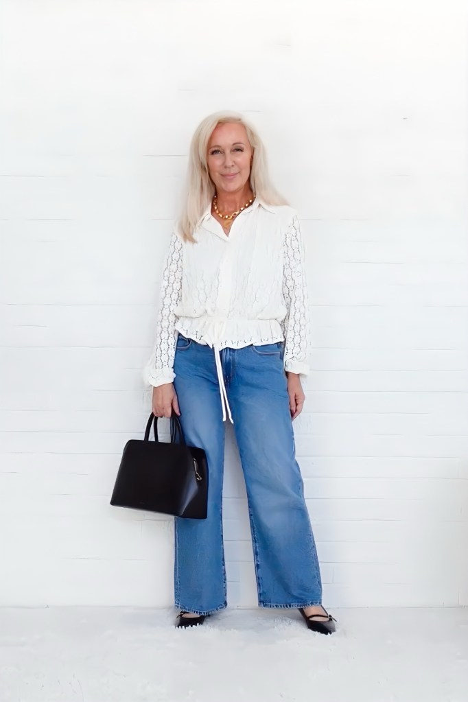 A woman with long, light-colored hair stands in front of a white wall. She is wearing a white blouse with lace details, light blue wide-leg jeans, and black flats. She holds a black handbag and has a necklace on.