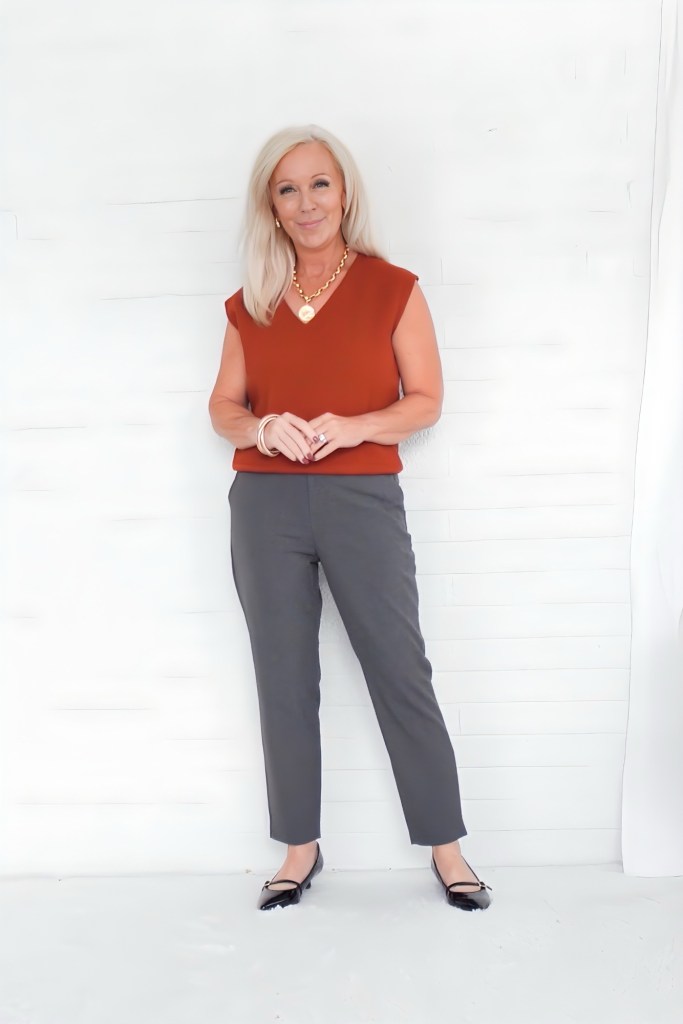 A woman with shoulder-length blonde hair stands confidently in front of a white background. She wears a rust-colored sleeveless top with gray pants and black flats. Her accessories include a gold necklace and bracelets.