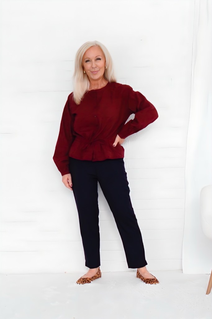 A woman with long blonde hair wearing a burgundy sweater and navy pants, standing confidently against a white background.