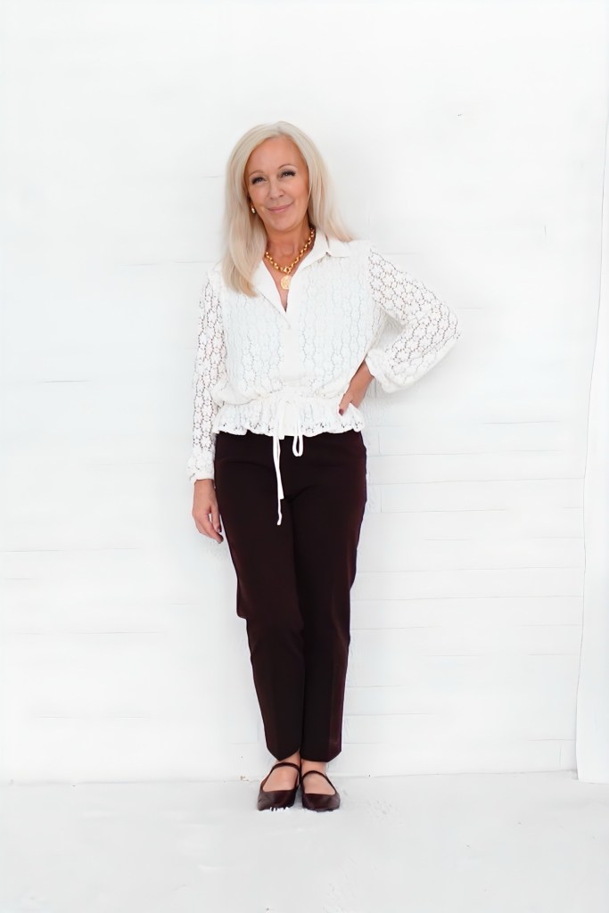 A woman with long, light-colored hair is wearing a white lace top and dark pants, standing against a white background.