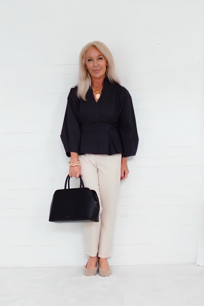 A woman with long blonde hair wearing a black blouse and light-colored pants, holding a black handbag, standing in front of a white wall.