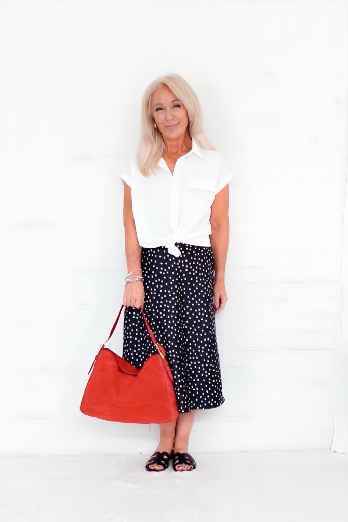 A woman stands in front of a white wall, wearing a white blouse tied at the waist and a black polka dot skirt. She holds a red bag and wears black sandals.