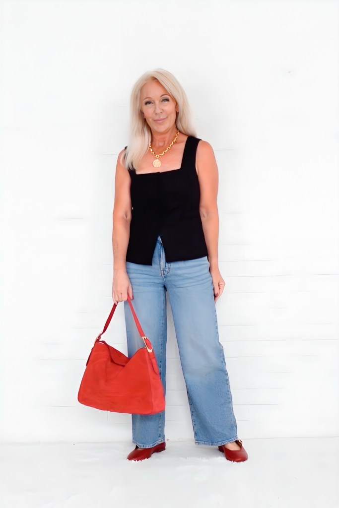 A woman wearing a black tank top and blue wide-leg jeans stands against a white background, holding a red bag.