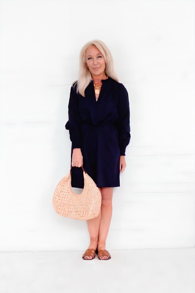 A woman stands in front of a white wall, wearing a navy dress and holding a woven bag. She has long blonde hair and is accessorized with a statement necklace.