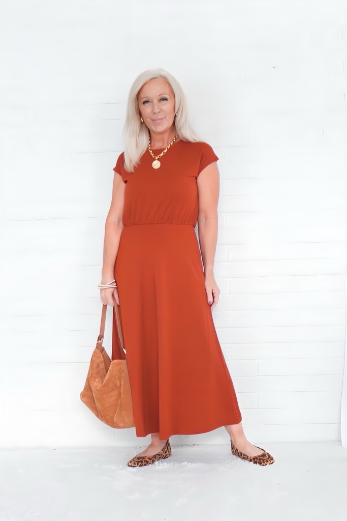 A woman wearing a rust-colored dress, accessorized with a gold necklace and a bag, stands in front of a white background.