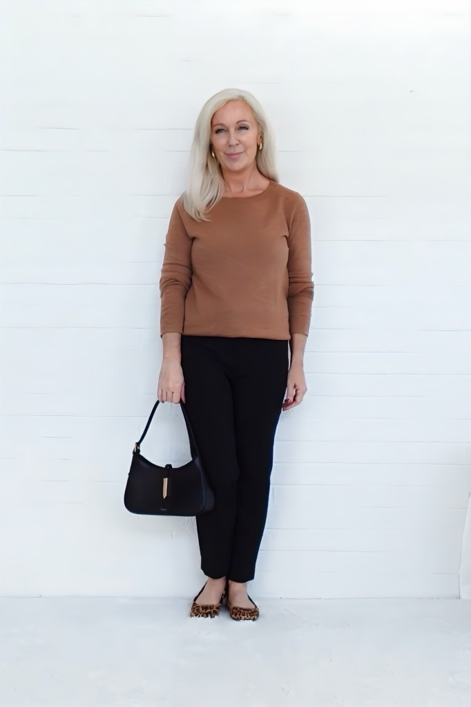 A woman stands against a white wall, wearing a brown long-sleeve top, black pants, and leopard print shoes. She holds a black handbag and smiles at the camera.