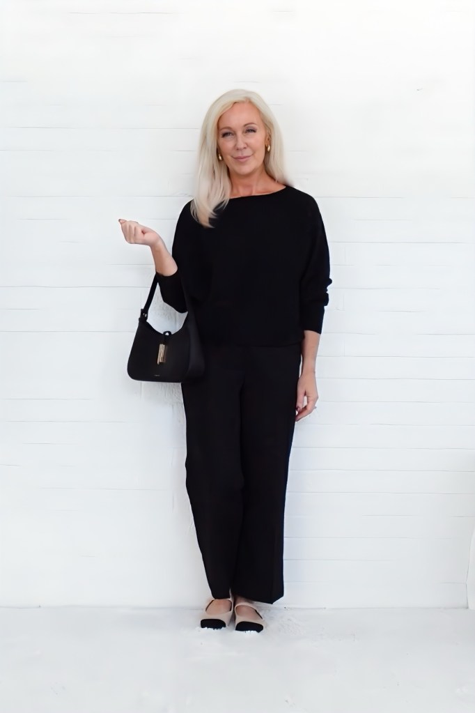 A woman standing against a white background, wearing a black sweater and black wide-leg pants, holding a black handbag and smiling.