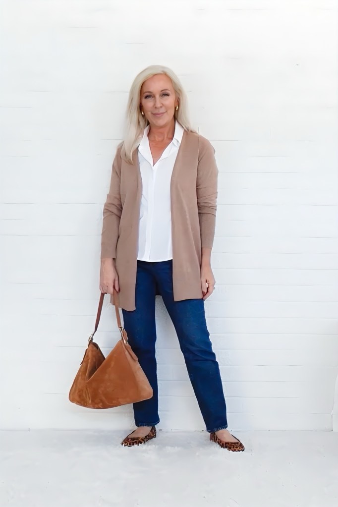 A woman with long, blonde hair poses in front of a white background, wearing a beige cardigan over a white blouse and blue jeans. She holds a brown handbag and has a confident smile.