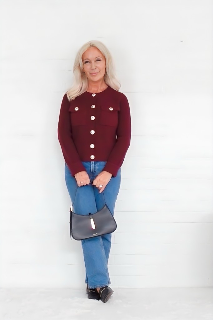 A woman with long blonde hair stands smiling against a white wall, wearing a burgundy cardigan with large buttons and blue jeans, holding a black handbag.