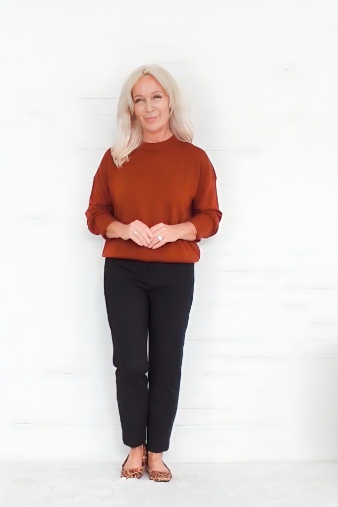 A woman stands smiling against a white background, wearing a rust-colored sweater, black pants, and leopard-print flats.